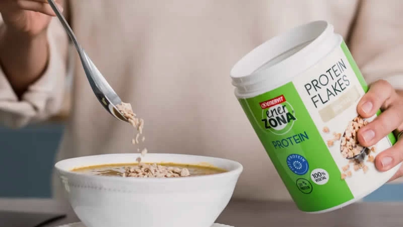 Una mujer echa ENER ZONA Protein flakes con una cuchara sobre un tazón de cereales